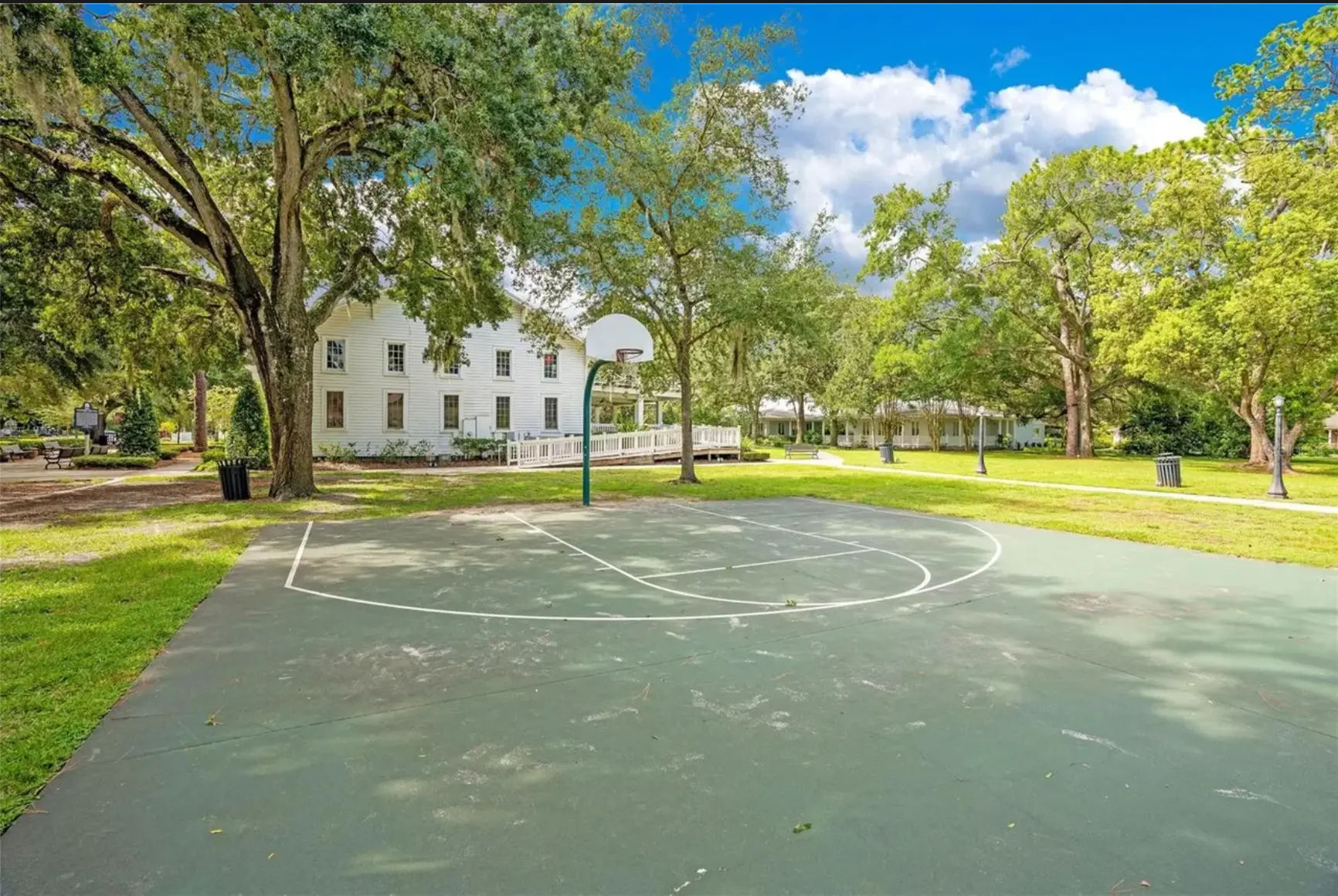 Community basketball court shaded by live oaks beside a historic white building